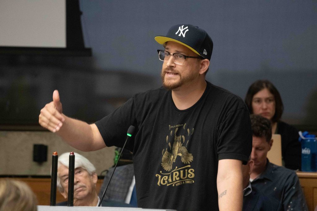 Image of Clifford Eiffler-Rodriguez in the foreground at a podium. He is wearing a Yankees hat and a black shirt with the logo of Icarus Wings and Things adorned on the front in gold.